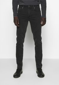 Jeans de mezclilla negra con un ajuste slim, que cuentan con cinco bolsillos, cierre de botón y detalles de costura a lo largo de las costuras. Combinados con zapatillas negras.