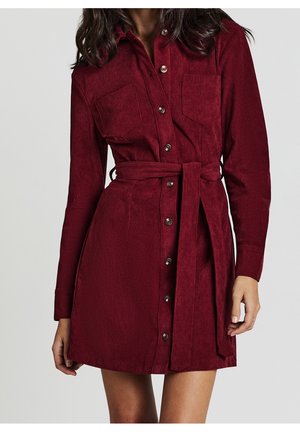 Femme portant une robe en velours côtelé bordeaux à manches longues, boutonnée, avec des poches poitrine et une ceinture nouée à la taille.