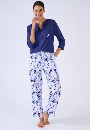 Donna sorridente con top blu a maniche lunghe con piccolo ricamo floreale e pantaloni larghi con motivo floreale, in piedi a piedi nudi contro sfondo bianco.