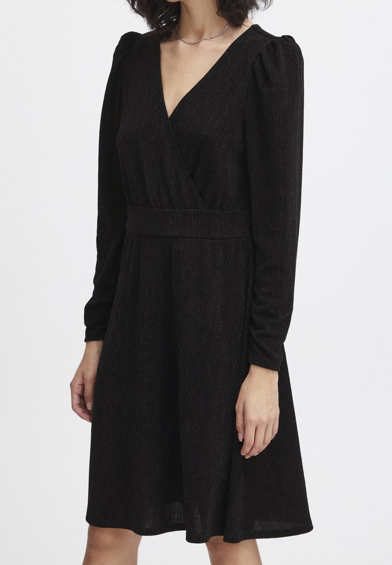 Femme portant une robe noire à manches longues, col en V, taille cintrée et épaules légèrement bouffantes sur un fond uni.