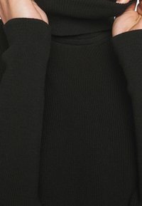 Pull en maille noire avec un col roulé haut. Le tissu présente une texture côtelée et des manches longues, créant une silhouette confortable.