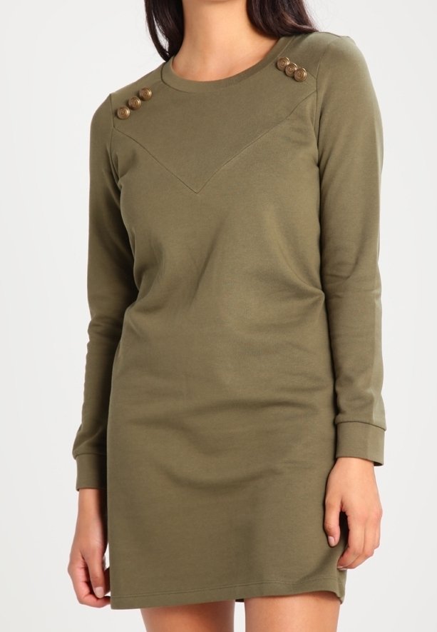 Femme portant une robe longue manches longues vert olive avec des détails de boutons sur les deux épaules, debout devant un fond uni.