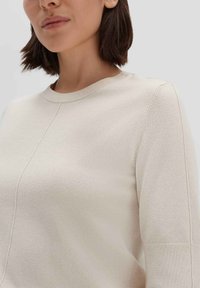 Helle beigefarbene Strickjacke mit Rundhalsauschnitt, die über feine Nähte und lange Ärmel verfügt. Weiche Textur, figurbetontes Design.