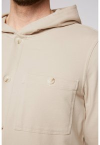 Sudadera con botones de color beige claro hecha de tejido texturizado. Cuenta con un bolsillo frontal, capucha y botones redondos, con detalles de costura visibles.