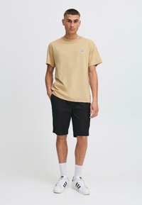 Camiseta de algodón beige con cuello redondo y mangas cortas, combinada con pantalones cortos negros. Zapatillas blancas con detalles negros y calcetines blancos.