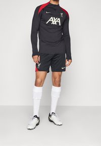 Schwarzes Langarmtrikot mit roten Akzenten, Logo und weißer Schrift, kombiniert mit schwarzen Shorts mit roten seitlichen Streifen und weißen Socken.