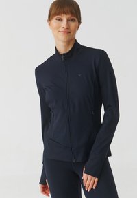 Mornarska majica za sport s patentnim zatvaračem, visokim ovratnikom, dugim rukavima, zatvorenim bočnim džepovima i prilagođenim krojem. Izrađena od glatkog, rastezljivog materijala.