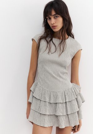 Ung kvinde med langt mørkt hår iført en ærmeløs, lysegrå ternet kjole med lagdelt flæse nederdel, poserer foran en hvid væg.