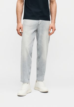 Mann trägt verblasste hellgraue Jeans, weiße Turnschuhe und ein schwarzes kurzärmeliges Hemd, steht vor einem schlichten Hintergrund.