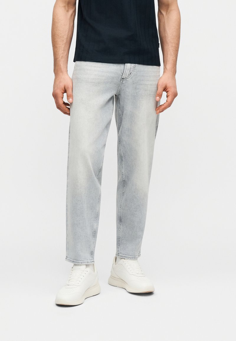 Άνδρας που φοράει ξεθωριασμένο ανοιχτό γκρι τζιν, λευκά αθλητικά παπούτσια και μαύρο κοντομάνικο πουκάμισο, στέκεται μπροστά σε απλό φόντο.