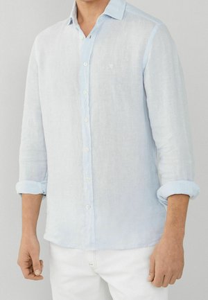 Homme portant une chemise bleu clair à manches longues boutonnée avec les manches retroussées et un pantalon blanc sur un fond neutre.