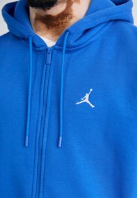 Blauwe rits-hoodie gemaakt van zachte stof; heeft koordjes en een wit Jumpman-logo op de linkerborst. Soepele textuur, casual ontwerp.