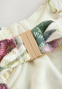 Bretea de sutien din dantelă florală cu cataramă aurie dreptunghiulară pe un fundal din plasă crem, evidențiind textura și contrastul culorilor.