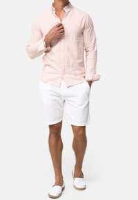 Mann trägt ein hellrosa gestreiftes Langarmhemd, weiße Shorts und weiße Slipper, steht mit einer Hand in der Tasche und der anderen Hand am Kragen.