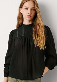 Blusa negra de manga larga con cuello alto, frente plisado y tejido sutilmente texturizado. Presenta costuras decorativas en el pecho.