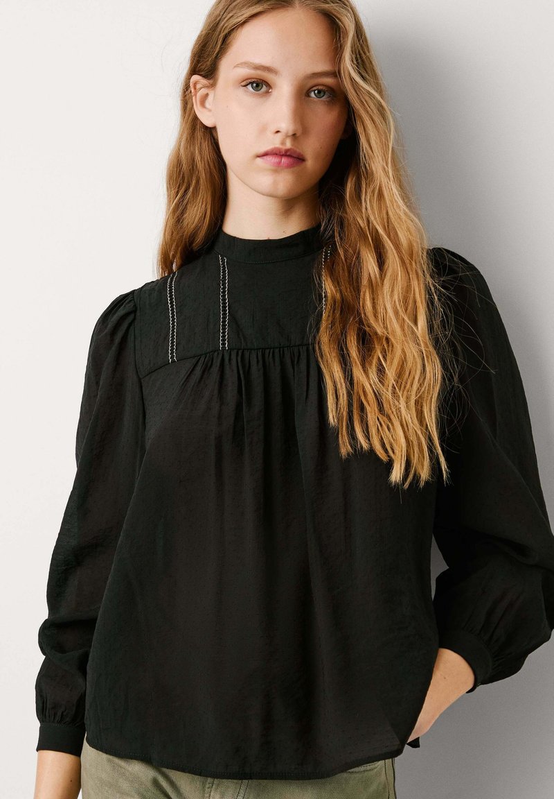Blusa negra de manga larga con cuello alto, frente plisado y tejido sutilmente texturizado. Presenta costuras decorativas en el pecho.