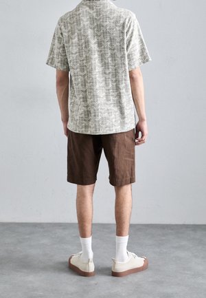 Mann steht mit dem Gesicht zu einer grauen Wand, trägt ein gemustertes Kurzarmhemd, braune Shorts, weiße Socken und weiße Sneaker mit braunen Sohlen.