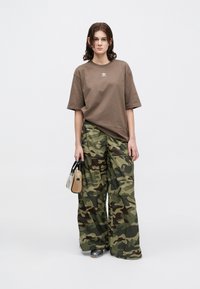 Tânără îmbrăcată cu un tricou Adidas maro, pantaloni largi din camuflaj, încălțăminte argintie, ținând o geantă transparentă cu un posetă maro în interior.