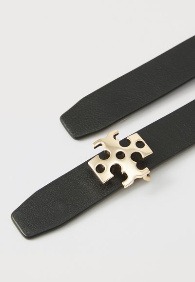 Černý kožený pásek se zlatým kovovým sponou s designem puzzle a kulatými výřezy. Plochý tvar s hladkým povrchem.