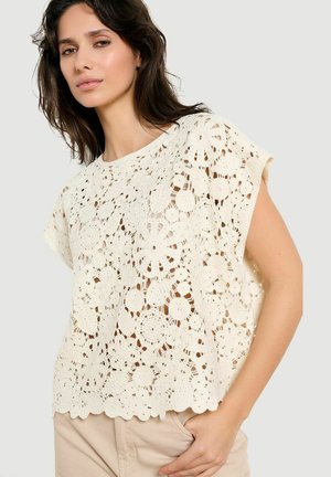 Vrouw draagt een kortärmelig crèmekleurig gehaakt kanten topje met bloemmotieven, gecombineerd met beige broek, staand tegen een effen achtergrond.