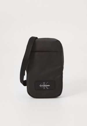 Petit sac bandoulière noir Calvin Klein avec poche avant et bandoulière en tissu réglable sur fond blanc uni.