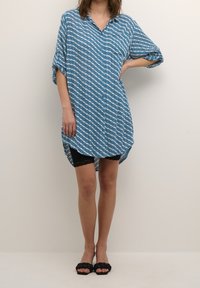 Robe bleu clair à boutons avec des motifs ondulés blancs, manches courtes et fentes sur les côtés. Associée à un short noir avec des bordures en dentelle et des sandales noires.