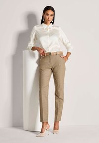 Witte satijnen blouse, beige gestreepte broek, accessorized met een riem en hooggehakte, nude schoenen. Model poseerde naast een cilindervormige voet.