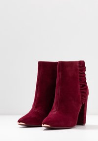 Stivaletti in suede borgogna con tacco a blocco alto, punta affusolata e dettaglio arricciato sul retro, dotati di accenti metallici sulla punta.