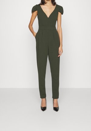 Olijfgroene jumpsuit met korte puffmouwen, V-hals, geplooide voorkant, taps toelopende pijpen en zijzakken, gecombineerd met zwarte hakken.
