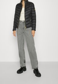 Schwarze gesteppte Pufferjacke mit hohem Kragen, goldenen Reißverschluss und Knöpfen, kombiniert mit grauen hoch taillierten Jeans und schwarzen Plateaustiefeletten.