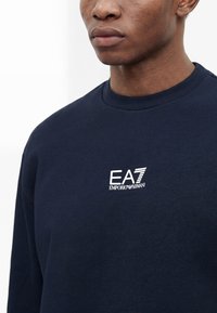 Άνδρας που φοράει σκούρο μπλε φούτερ με λευκό λογότυπο EA7 Emporio Armani στο στήθος, εμφανίζεται από τον λαιμό έως το μέσο του κορμού.