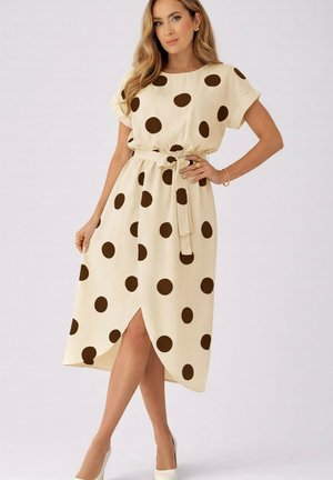 Femme portant une robe midi crème à grands pois marron, manches courtes, taille ceinturée et fente devant, posant avec la main sur la hanche.