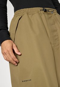 Beige bukse med elastisk midje og glatt tekstur, dekorert med folder. Har snøring og "OAKLEY" merkevare på nedre del av beinet.