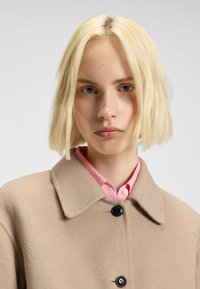 Beige wool coat with a collar, single black button closure, worn over a light pink collared shirt. Smooth texture, simple design.