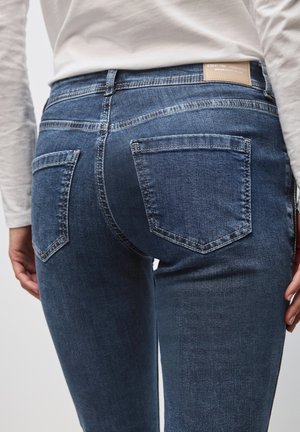 Persona vistiendo un pantalón de mezclilla azul y una camisa de manga larga blanca, con bolsillos traseros visibles y detalles de costura en los pantalones.