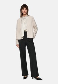 Hellbeige Strickjacke mit Rippstruktur, mit Knopfverschluss, kombiniert mit einer weißen Bluse mit Schleife, schwarzen weiten Hosen und schwarzen spitzen Schuhen.