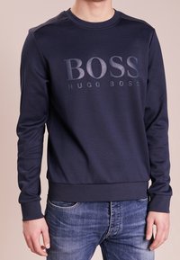 Námořnická modrá mikina vyrobená z bavlněné směsi, která má na přední straně vyvýšené logo "BOSS HUGO BOSS", žebrované manžety a pas.