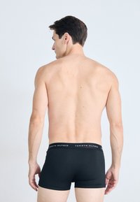 Man facing away, shirtless, wearing black boxer briefs with Tommy Hilfiger logo on waistband against plain background.