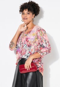 Mehrfarbige, mit abstrakten Mustern gestaltete Bluse mit V-Ausschnitt und Dreiviertelärmel. Kombiniert mit einer glänzenden roten Clutch und einem schwarzen Lederrock.