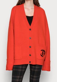 Czerwony sweter z dzianiny, z dekoltem w serek, w oversize'owym fasonie, z czarnymi guzikami, dwiema przednimi kieszeniami i wyszytym logo na lewej kieszeni.