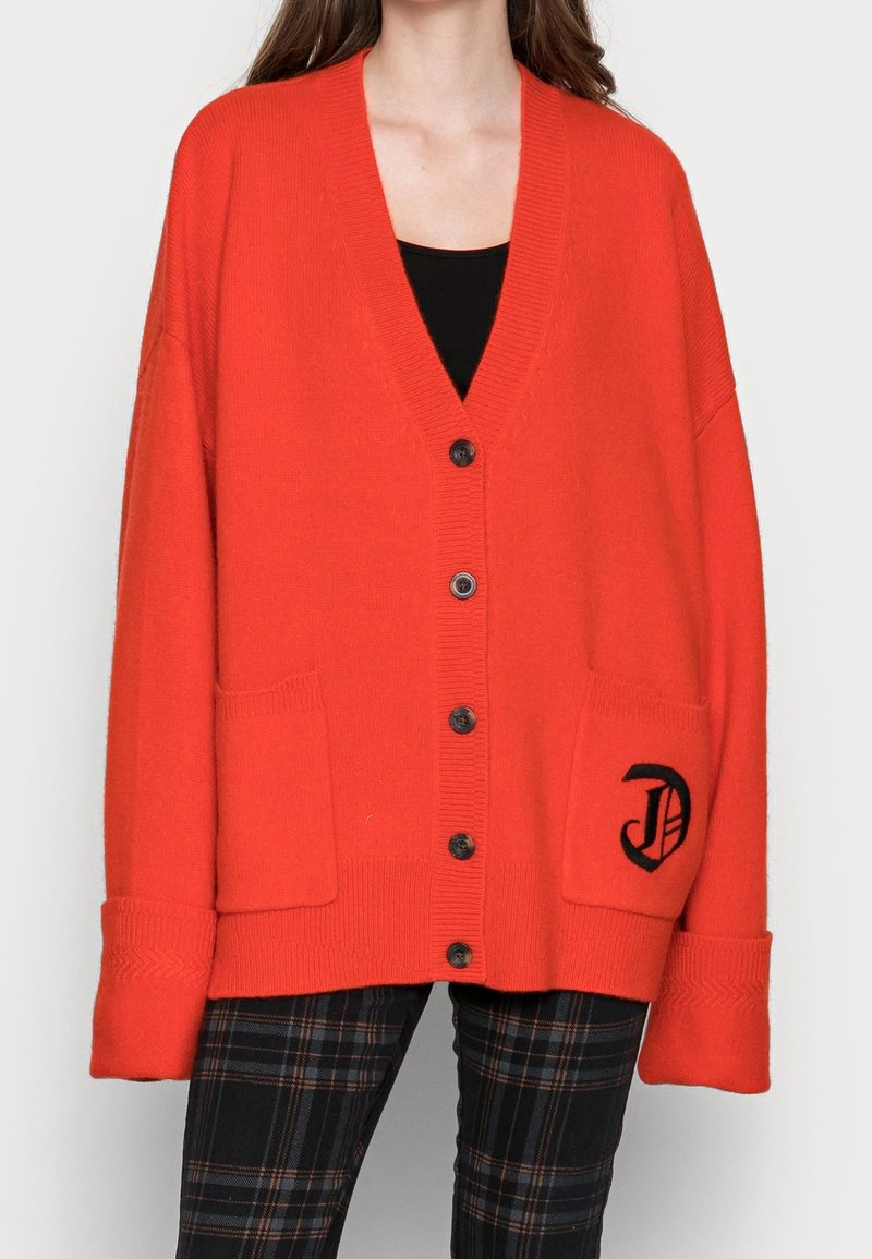 Czerwony sweter z dzianiny, z dekoltem w serek, w oversize'owym fasonie, z czarnymi guzikami, dwiema przednimi kieszeniami i wyszytym logo na lewej kieszeni.