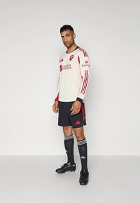 Tenue de football à manches longues blanche avec des accents rouges, shorts noirs, chaussettes noires à rayures blanches et crampons noirs. Ornée de logos de sponsors.