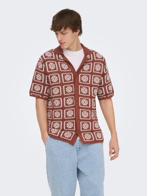 Jeune homme portant une chemise marron et blanche à manches courtes à motifs, boutonnée sur un t-shirt blanc et un jean bleu clair, les mains dans les poches.