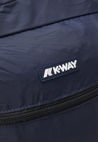 Navy blue bag fabric with a smooth texture, featuring a visible black zipper and a white logo label stitched on a dark blue patch.