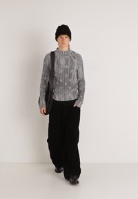 Γκρι ριγωτό πουλόβερ με αστέρια, μαύρο φαρδύ παντελόνι, μαύρο σκουφί και τσάντα ώμου. Το μοντέλο περπατά σε ανοιχτό φόντο.