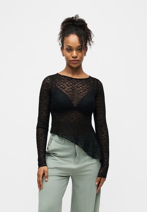 Femme aux cheveux bouclés portant un haut transparent en dentelle noire à manches longues par-dessus un sous-vêtement noir et un pantalon taille haute gris clair.