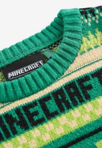 Grüner gestrickter Pullover mit einem gerippten Kragen, der ein Minecraft-Logoetikett und ein gemustertes Design in verschiedenen Grüntönen und Creme zeigt.