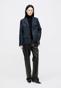 Donna che indossa un blazer in denim scuro con bottoni dorati, dolcevita nera, pantaloni in pelle neri e scarpe nere a punta con punte dorate.