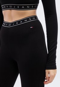 Μαύρο ριμπ κολάν με φαρδιά μαύρη ζώνη που φέρει λευκό κείμενο "TOMMY JEANS", μικρή τρίχρωμη λεπτομέρεια στο πλάι, λεία υφή.