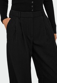 Sorte bukser med knapper og stripe, høyt liv, folder, sidelommer og teksturert stoff.